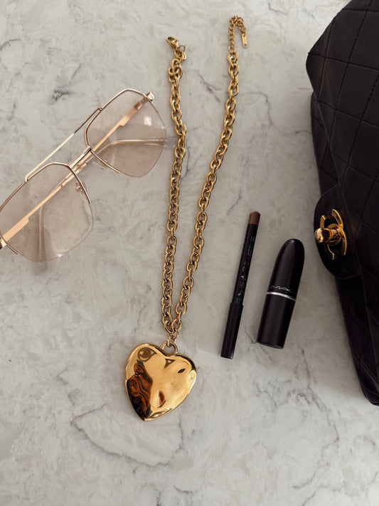 Gold heart-shaped necklace, sunglasses, makeup items, and a black handbag on a marble surface