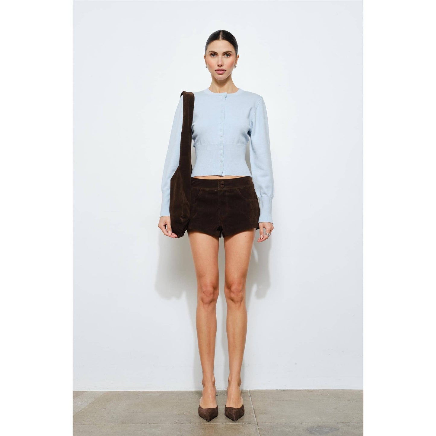 Woman wearing a light blue knitted cardigan and brown suede shorts, accessorized with a brown bag.