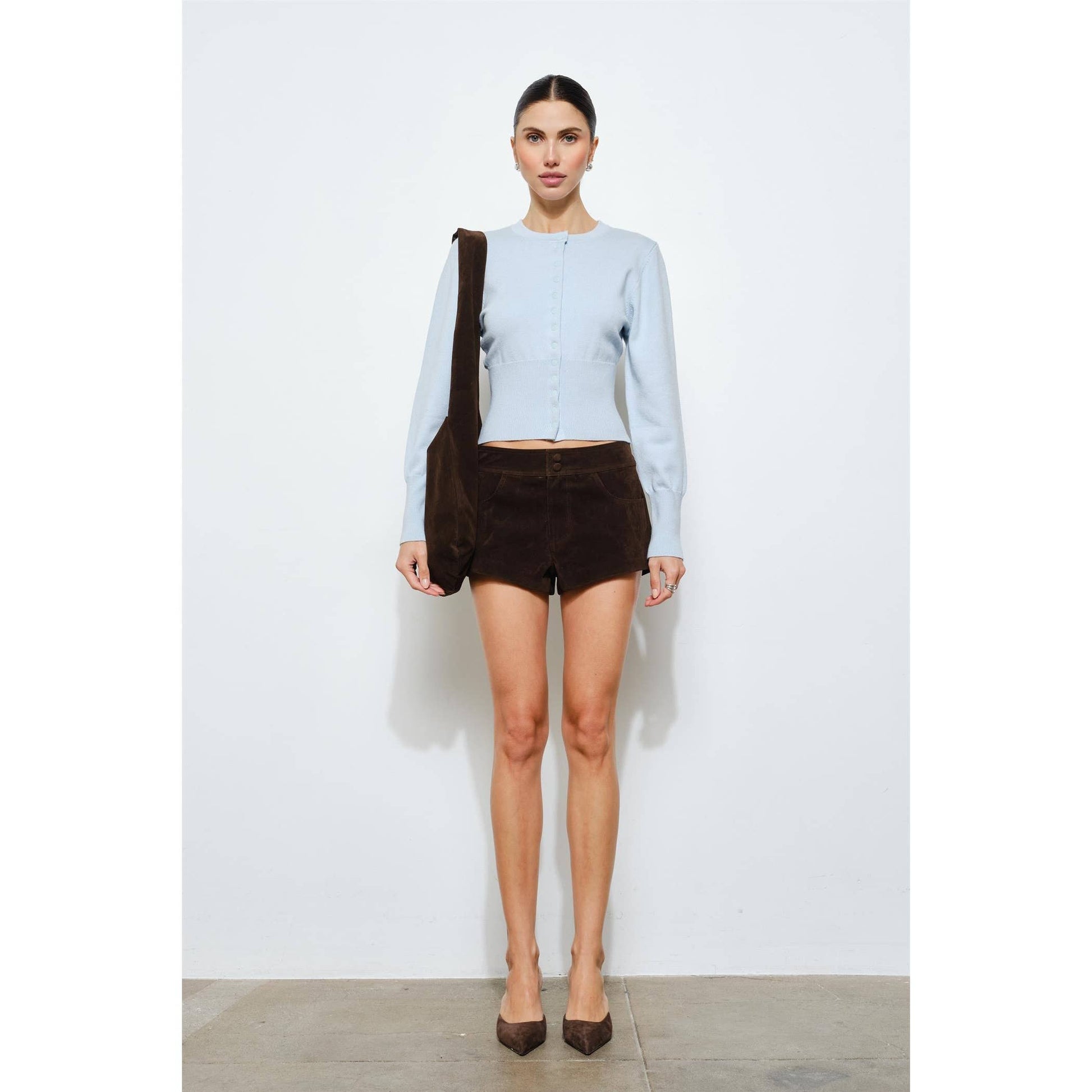 Woman wearing a light blue knitted cardigan and brown suede shorts, accessorized with a brown bag.